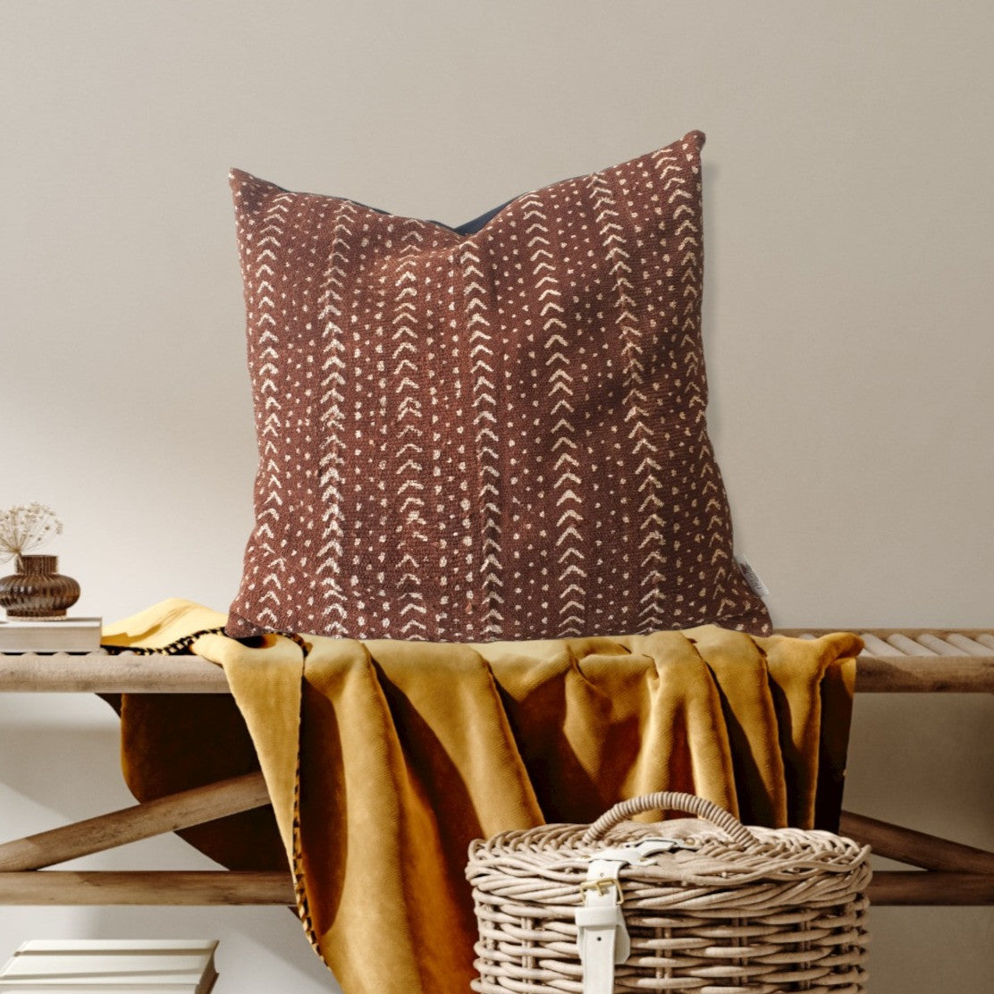 Decorative pillow with brown and beige pattern on a wooden bench with a yellow blanket and wicker basket.