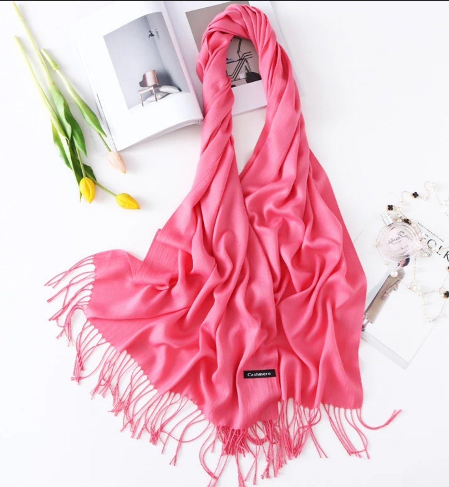 Pink scarf with tassels on a white surface with a magazine and flowers.