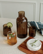 Brown carafe with glasses of iced tea on a white surface with a wooden board and cheese.