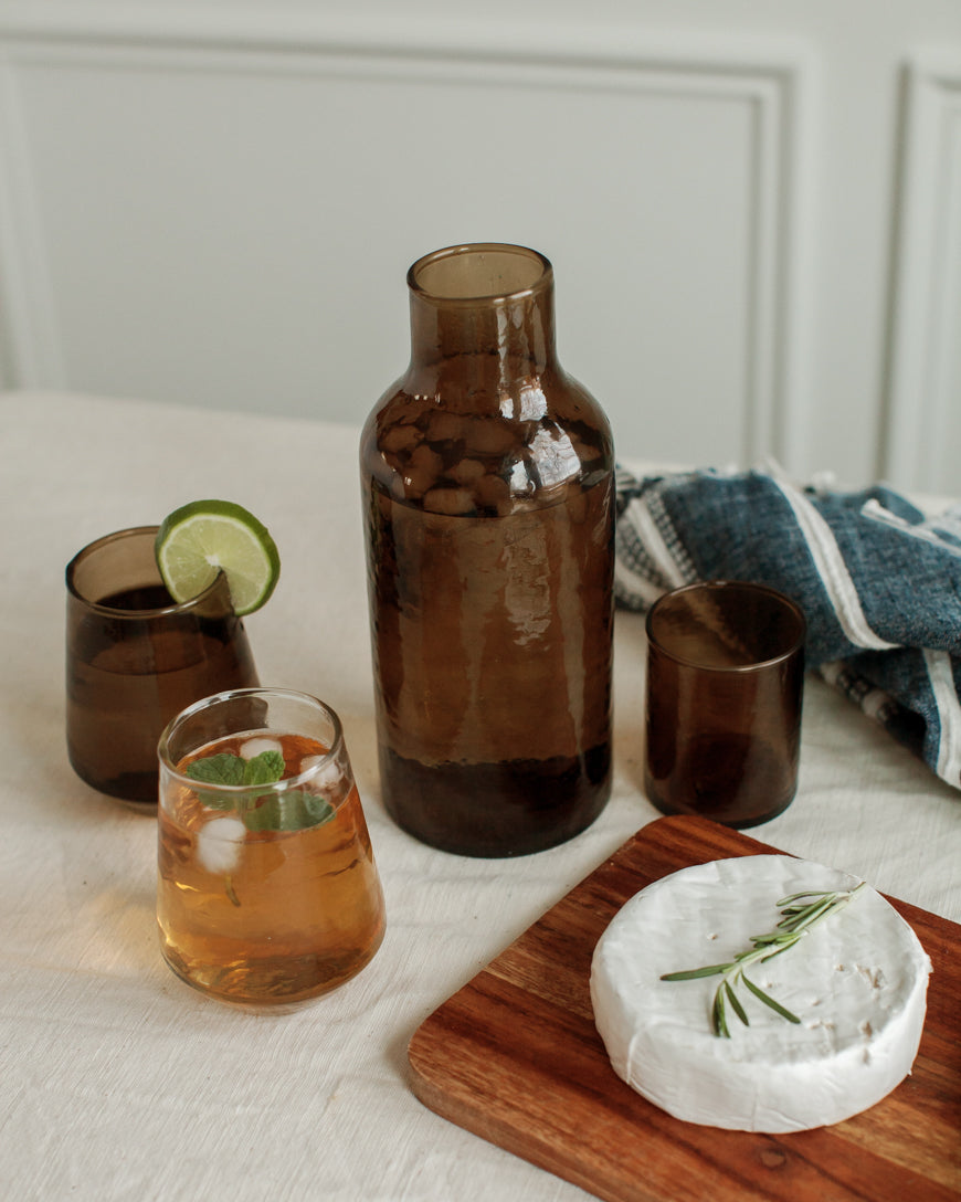 Brown carafe with glasses of iced tea on a white surface with a wooden board and cheese.