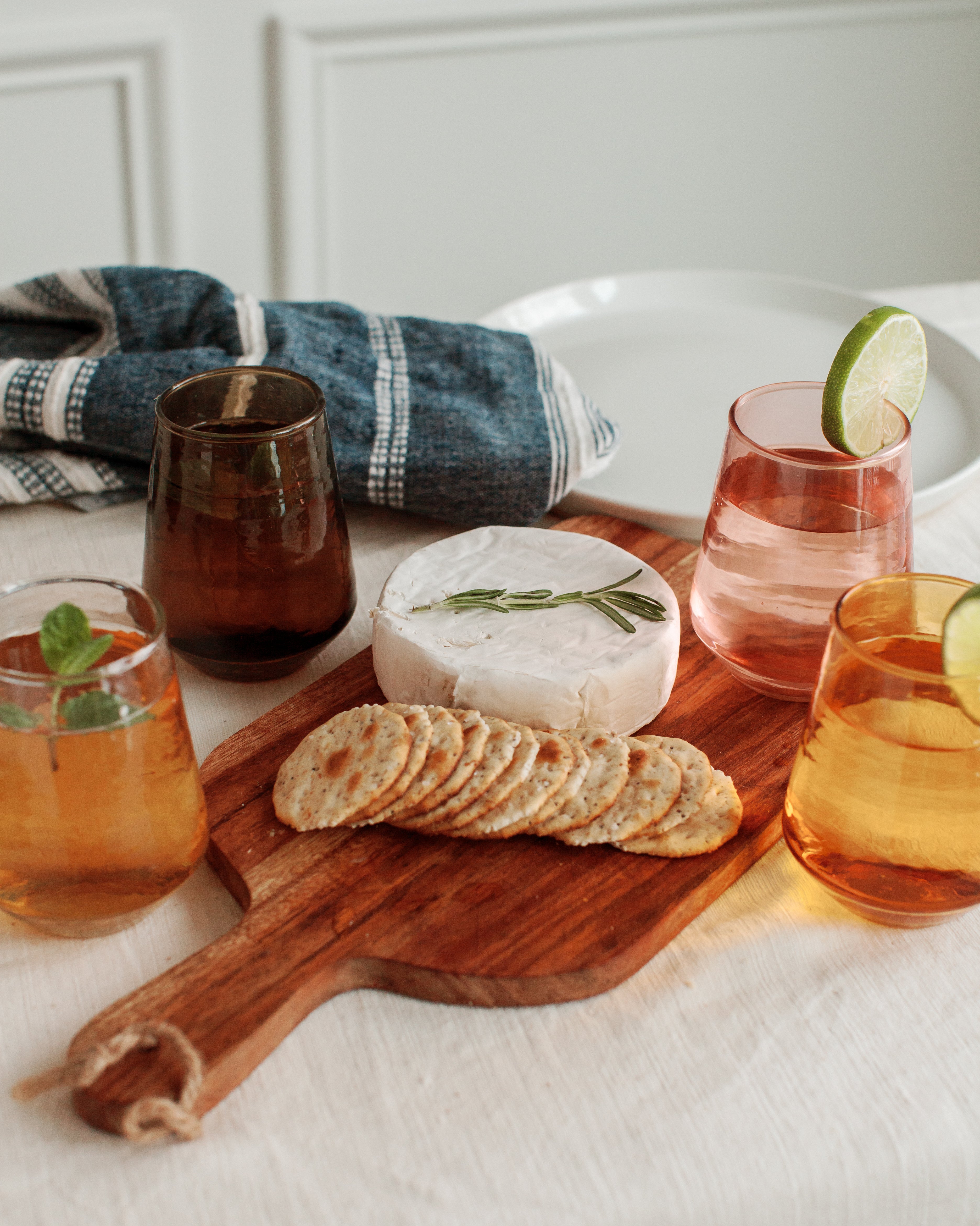 Wooden cutting board with cheese, crackers, and glasses of tea on a white surface.