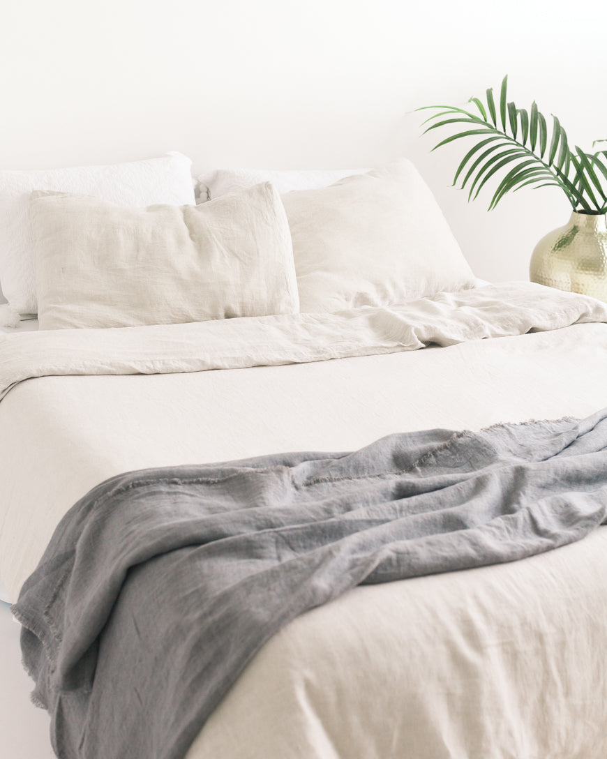 Neatly made bed with white bedding and a gray throw blanket, plant in the background.